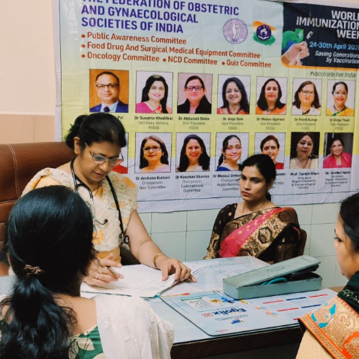 Gynecologist consulting women patients during a FOGSI World Immunization Week 2026 awareness program in a hospital, with an educational vaccination campaign banner displayed in the background.